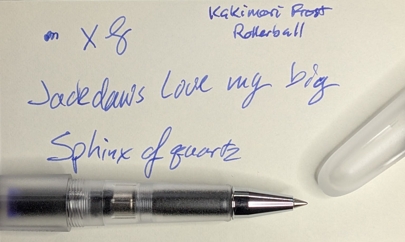 Photo of a rollerball pen with a transparent but frosted body and cap. On the paper is written "Kakimori Frost Rollerball", "Jackdaws love my big sphinx of quartz", and some scribbles, all in blue-purple ink.
