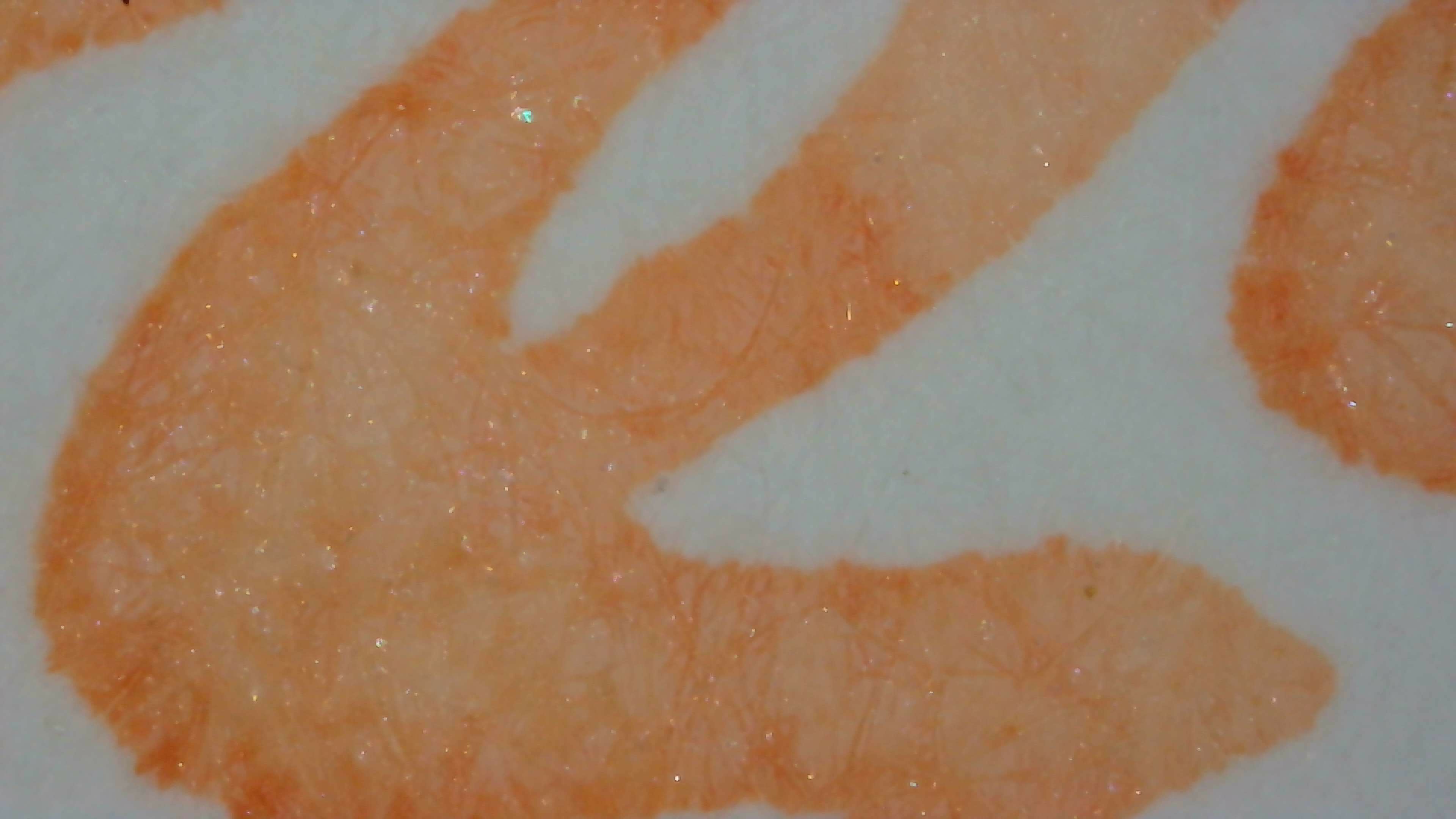 Photo of ink on paper under magnification on Iroful paper. The ink is peach colored and very light. The edges of the lines are very smooth, and on the left side they are a little darker. In the upper part of a loop in the line a section is even more pale. There is a light amount of visible shimmer particles evenly distributed in the ink.