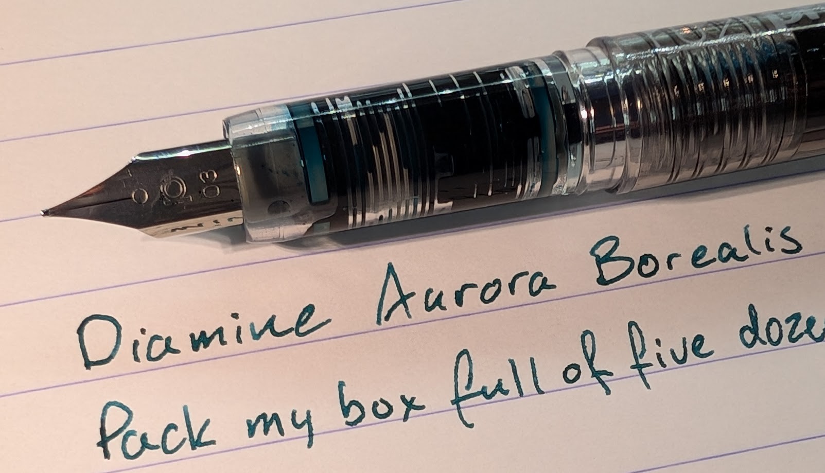 Photo of a fountain pen and writing sample. Fountain pen is a Platinum Preppy with a Black body. Only a small bit of the body is visible. The nib is marked "03" as it's a Fine nib. There is ink in the transparent section. The ink is Diamine Aurora Borealis, which is a medium blue/green color. The writing shows some shading, darker areas where more ink has settled. The visible portion of the writing sample reads: "Diamine Aurora Borealis Pack my box full of five doze"