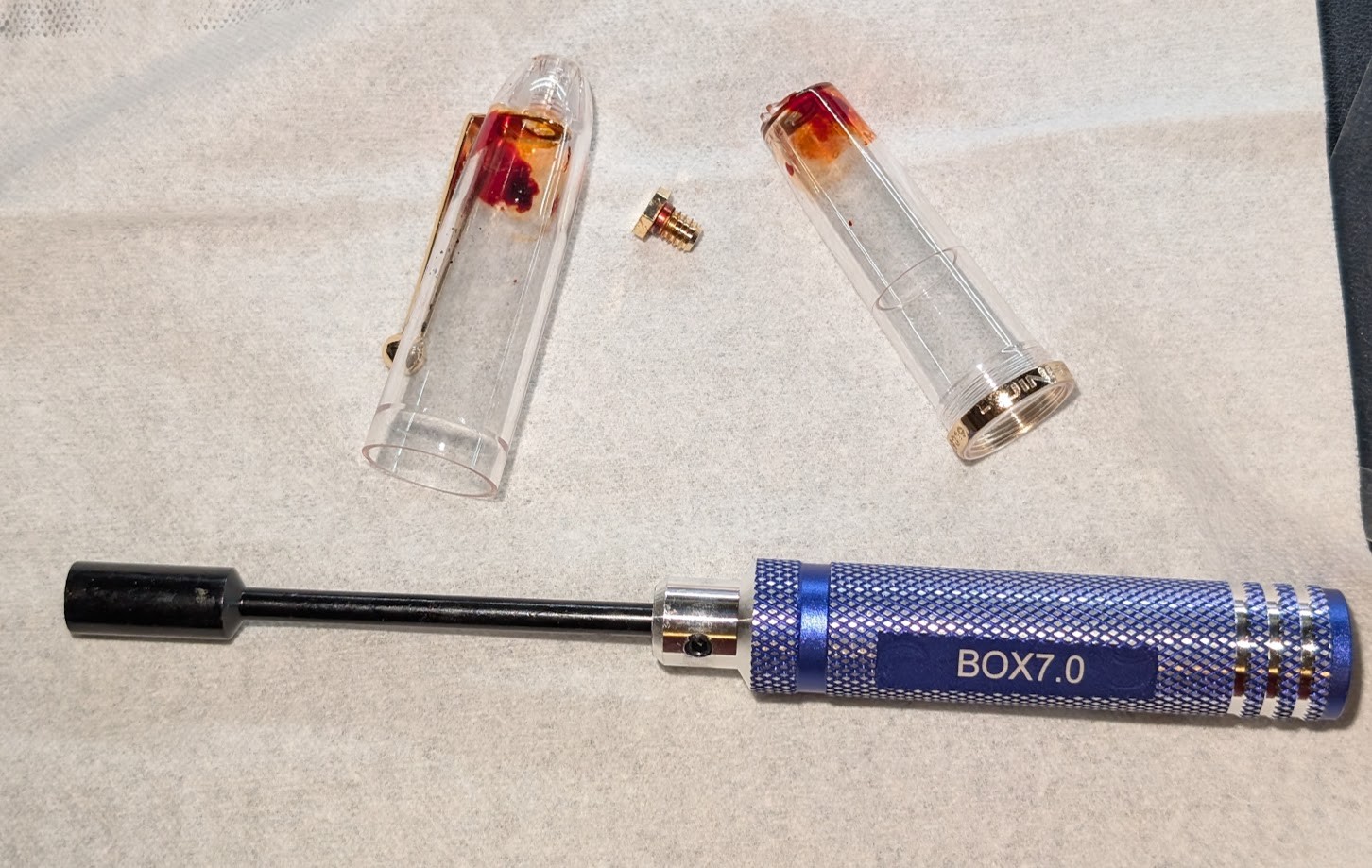 Photo of several items spread out on a tissue paper: - A blue nut driver with a long handle and shaft. It is labeled "BOX7.0" - The Jinhao 9019 outer cap with the clip attached and red ink near the tip. - The Jinhao 9019 inner cap with red ink near the tip - The screw that holds the inner cap, outer cap, and clip together. It is only a few mm long and has a 7mm hex head on it.