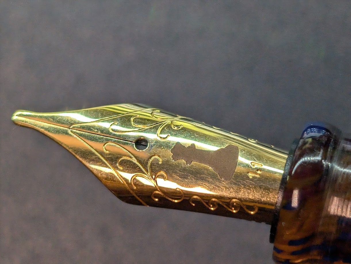 Photo of a fountain pen nib. The nib is yellow in color but is not gold. The tip of the nib is flat with rounded corners (stub). There is an outline near the edge of the nib. Inside that are some engraved curving lines which branch off in various swirls. In the center is the breather hole and underneath that is a dark engraving of a chess piece which appears to be a queen. Under the logo is stamped "1.1" indicating the nib size as 1.1mm.