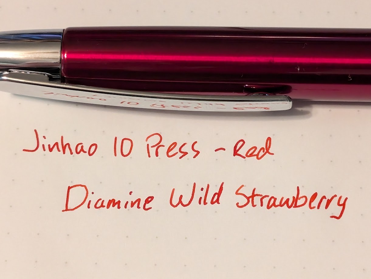 Photo of a fountain pen above a writing sample on a piece of paper. The fountain pen is dark red and shiny with a silver tip and silver clip. The writing sample is in bright red ink and reads: Jinhao 10 Press - Red Diamine Wild Strawberry