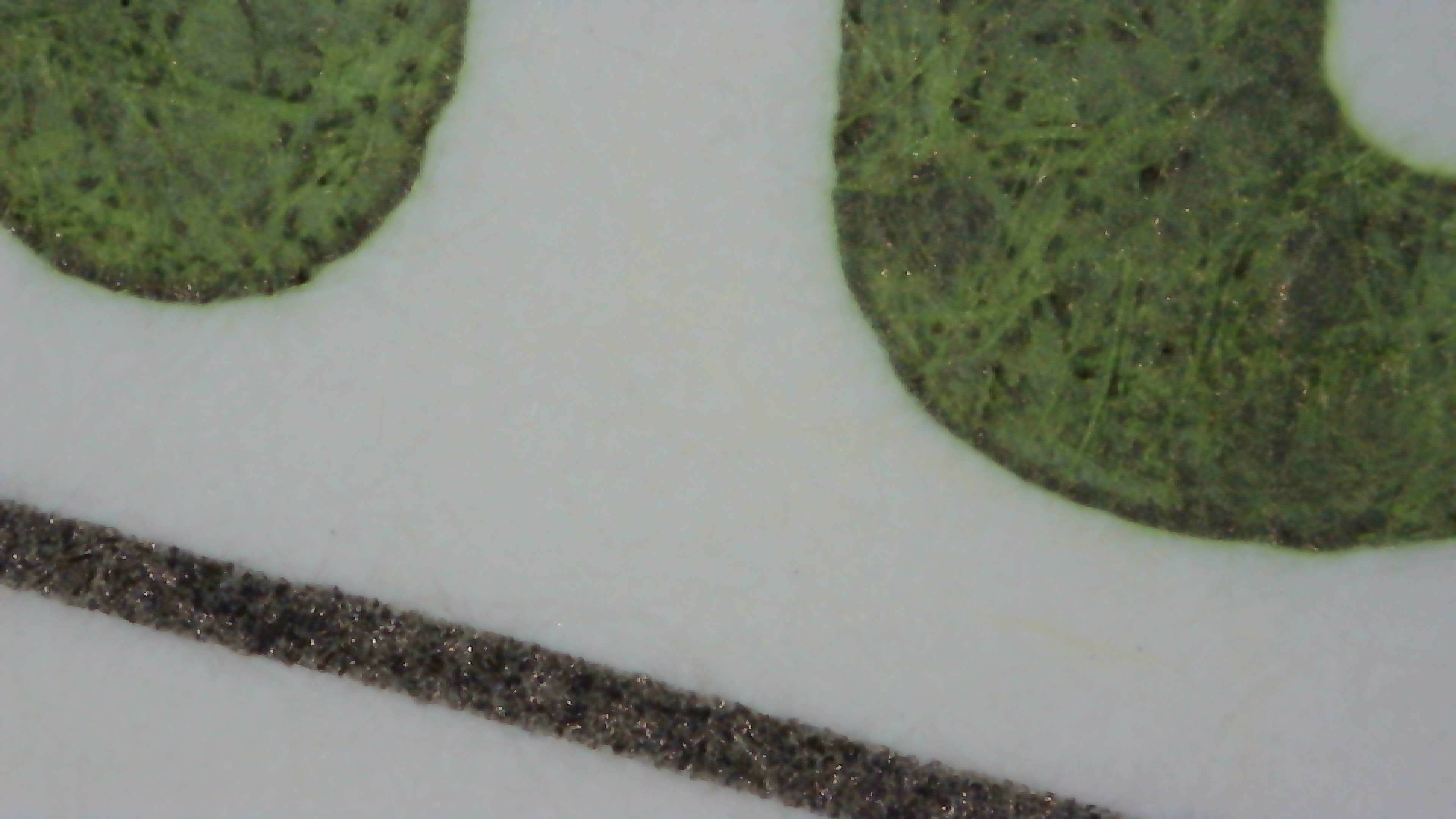 Photo of the ink on a Iroful card under high magnification. Paper fibers are visible. Most of the image consists of olive green ink with some spots which are darker. The edges of the lines are smooth.