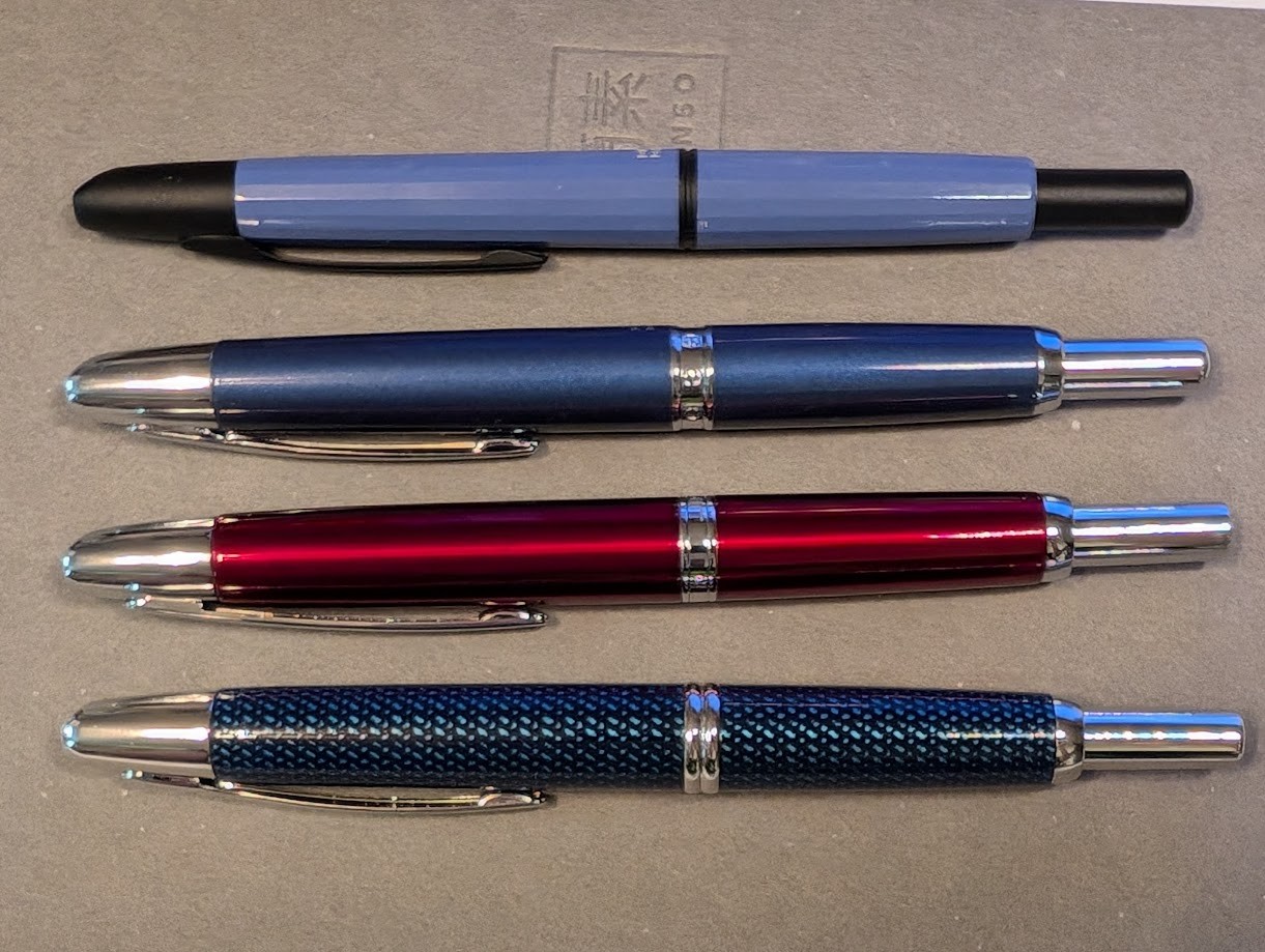 Photo of four fountain pens, placed horizontally and fully visible. From top to bottom they are: * Majohn A2 which is a flat blue-gray color with black trim and a faceted surface. * Majohn A1 in a dark shiny metallic blue color with silver trim and smooth surface. * Jinhao 10 Press in dark shiny red color with silver trim. * Pilot Vanishing Point in Blue Carbonesque pattern which is dark blue with a repeating pattern of lighter blue hashed-like splotches. The surface is slightly textured. It has silver trim. Each pen has slightly different trim in various ways, the shapes are a little different than one another but still similar.