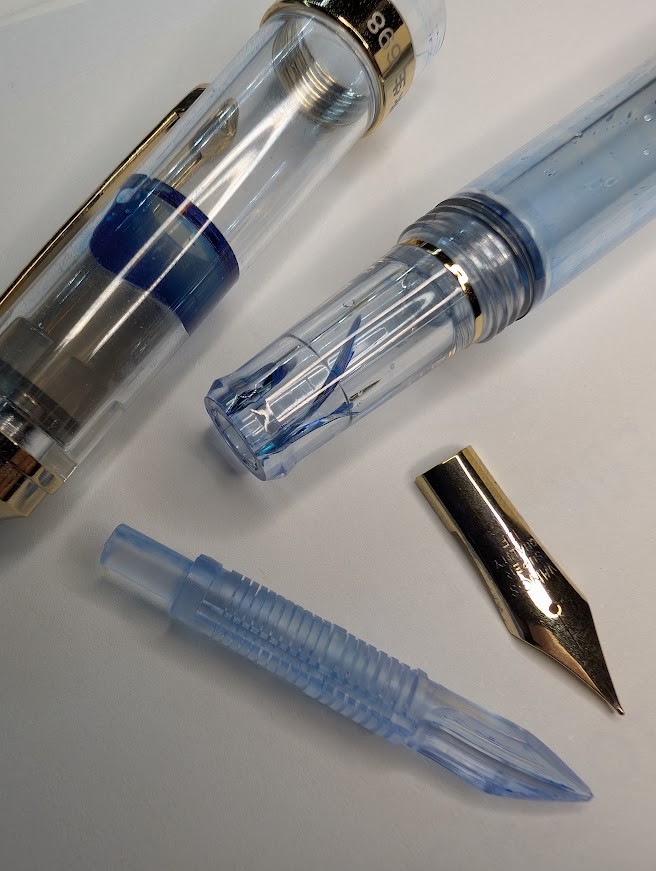 Photo of a partially disassembled fountain pen. The body and cap of the pen are clear with gold accents. The nib and feed and laying separately at the bottom of the photo. The body of the pen is in the top right. There are large cracks in the section that radiate out from the tip. The cap is laying at the top left of the photo. There is blue ink inside the cap between the cap liner and the outer cap.
