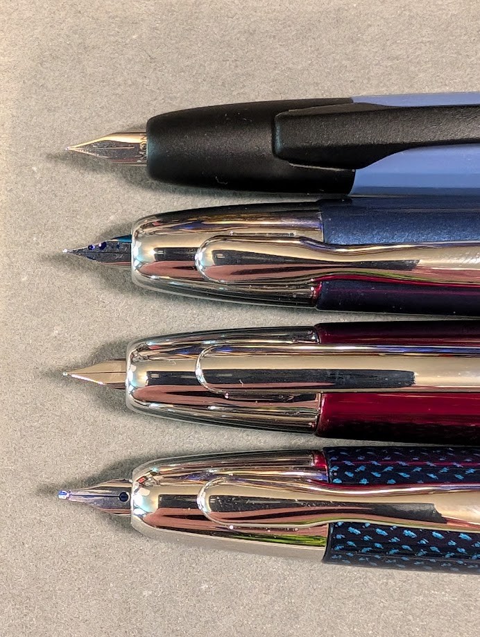 Photo of four fountain pens, placed horizontally and partially visible. Photo is focused on the tips of the pens with the nibs extended. From top to bottom they are: * Majohn A2 which is a flat blue-gray color with black trim and a faceted surface. The nib is silver and very pointy and extends slightly farther than the other pens. * Majohn A1 in a dark shiny metallic blue color with silver trim and smooth surface. The nib is silver and pointy and has a few splotches of blue ink on it. * Jinhao 10 Press in dark shiny red color with silver trim. The nib is silver and pointy, but clean, other than the ink it's approximately the same as the A1. * Pilot Vanishing Point in Blue Carbonesque pattern which is dark blue with a repeating pattern of lighter blue hashed-like splotches. The surface is slightly textured. It has silver trim. The nib looks significantly different to the others, it has a stub end and a large visible breather hole.