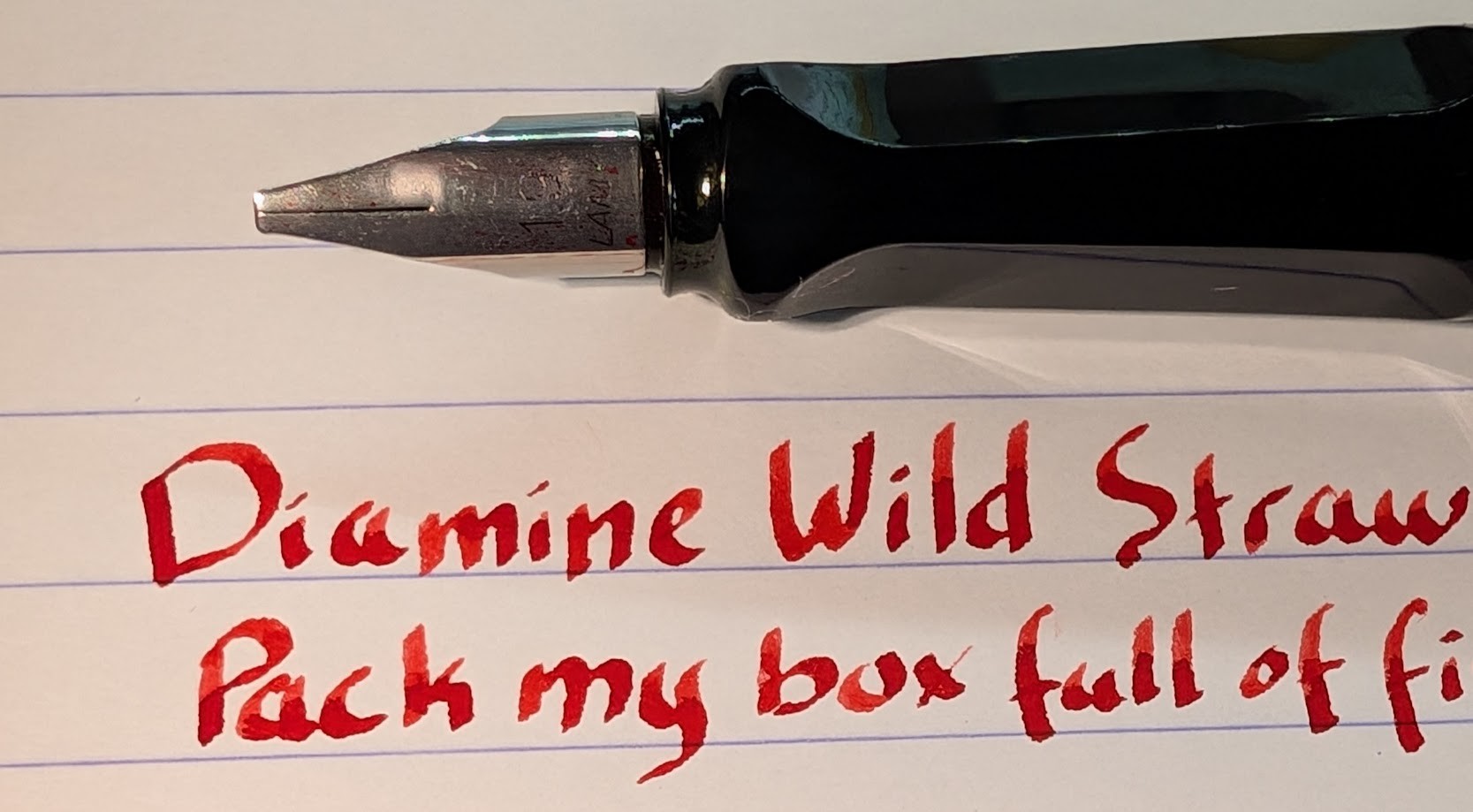 Photo of a fountain pen and writing sample. Fountain pen is a Lamy Joy with a Black section. The nib is marked "1.9 LAMY" as it's a 1.9mm stub nib. The ink is Diamine Wild Strawberry, which is a bright red color. The writing shows some shading, darker areas where more ink has settled. The visible portion of the writing sample reads: "Diamine Wild Straw Pack my box full of fi"