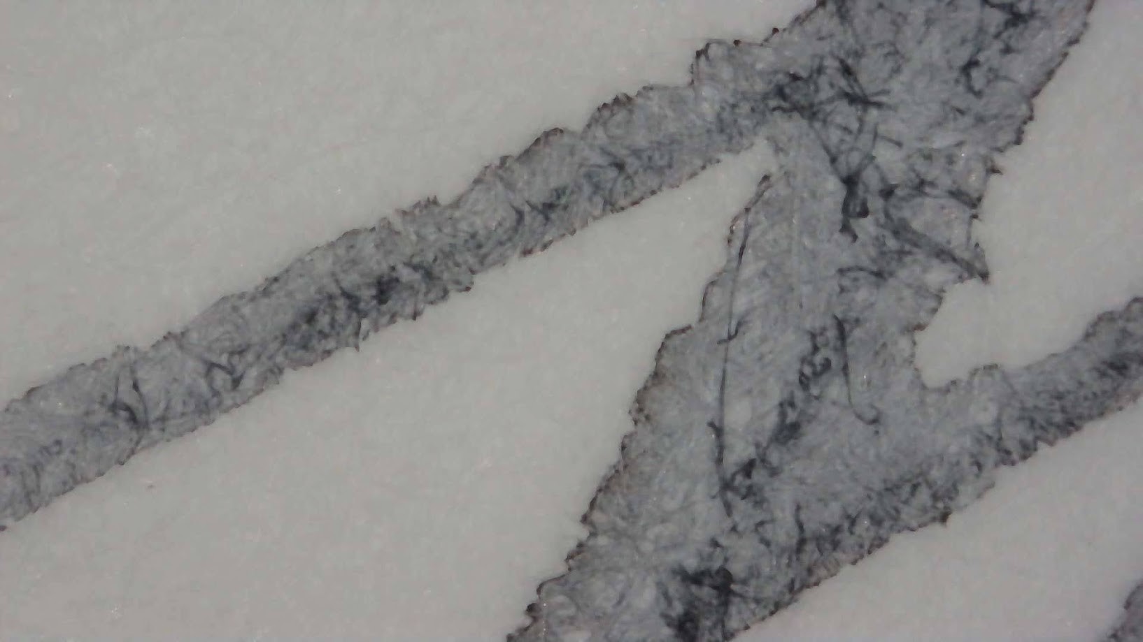 Photo of ink writing under a microscope. Paper fibers are visible. The ink is mostly light gray with some dark gray around the edge and in the center where ink has pooled. The general shape of the writing is a zig-zag but no distinct letters are visible.