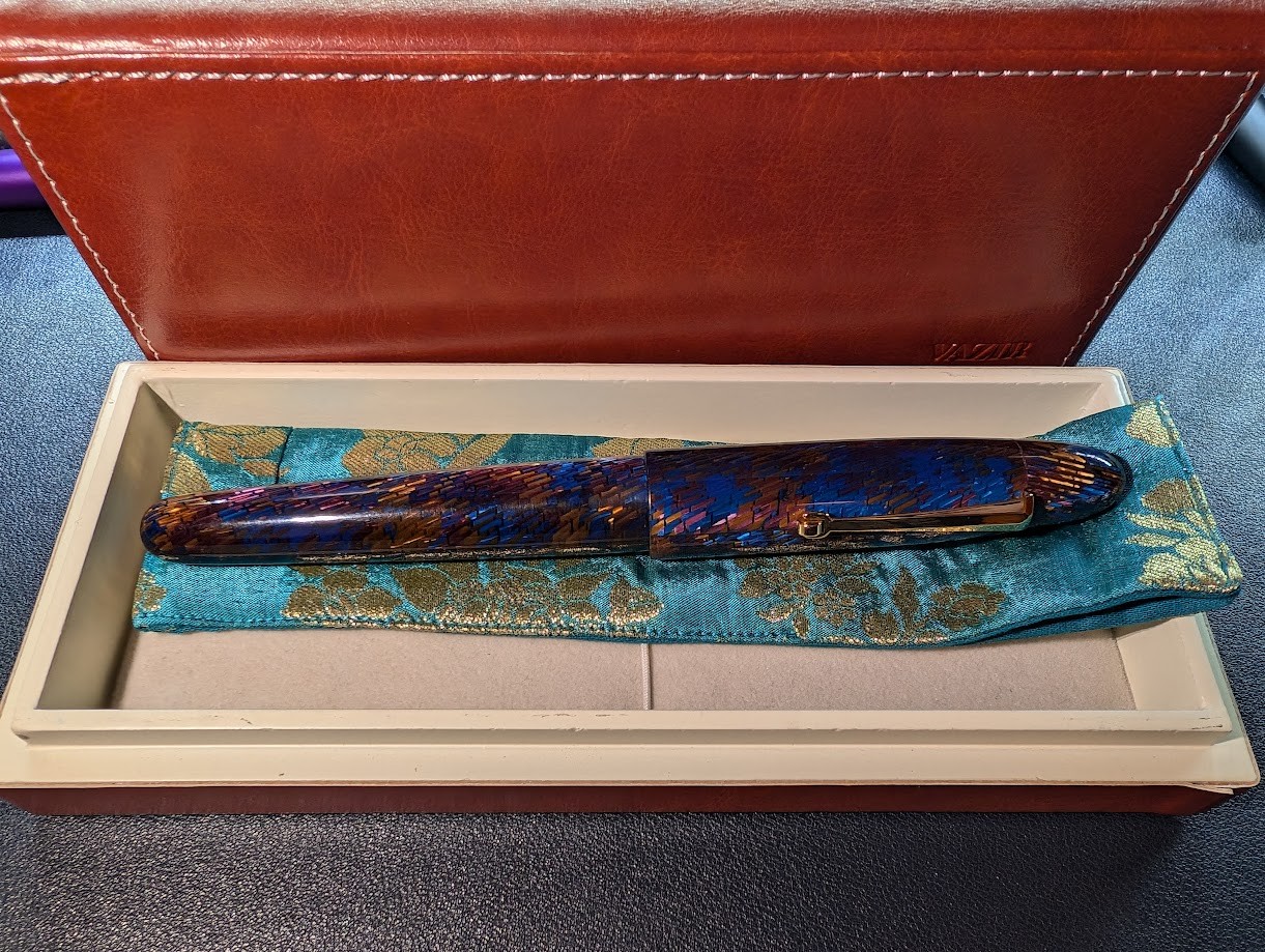 Photo of the Vazir Meteor Shower pen in a case. The pen is primarily blue with angled rows of material under the surface which catch the light in different colors including dark blues, yellows, and reds. The pen is sitting on top of a fabric pen pouch which is light blue with a flowery design in yellow thread. Under that is a padded bottom with an elastic strap. The gift box in which the pen sits is some sort of hardboard/soft wood like MDF. It is light brown inside. The outside of the box appears to be coating in brown leather-like material with stitching around the edges. The Vazir name is visible in the bottom right corner.