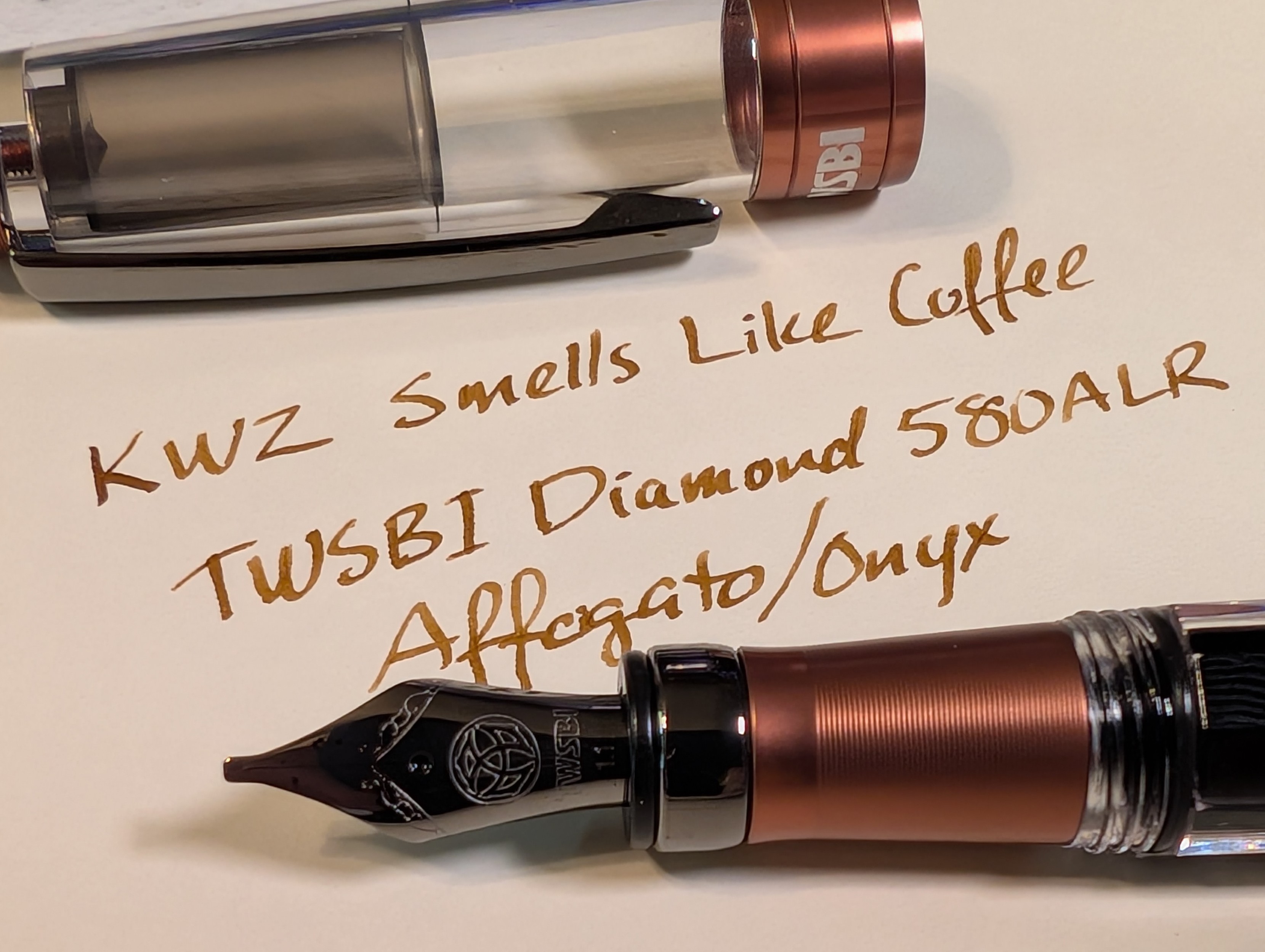 Photo of a fountain pen and writing sample on a piece of paper. The pen cap is removed and sitting across the top. The nib end of the pen is laying across the bottom. The pen is is clear plastic with metallic medium brown accents. The nib and clip are dark gray, almost black. The writing sample reads "KWZ Smells Like Coffee / TWSBI Diamond 580ALR Affogato/Onyx" in medium brown ink.