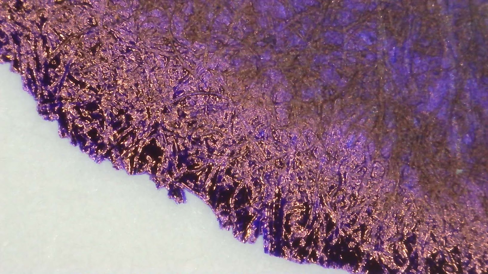 Photo of a bluish purple ink blob under magnification. Paper fibers are visible. Near the edge the ink is reflective and shiny red.