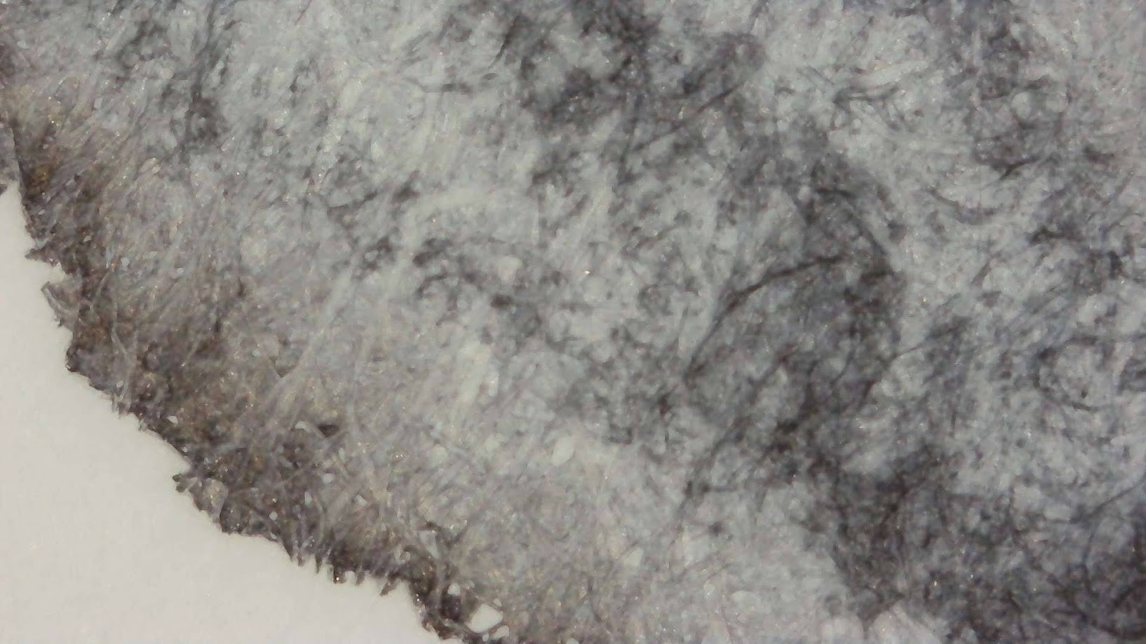 Photo of ink blob on paper under a microscope. Paper fibers are visible. The ink is mostly light gray with very dark gray around the edge and medium gray in the center where ink has pooled.