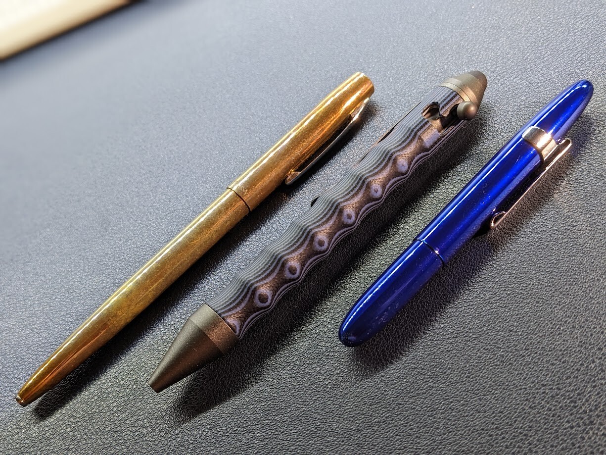 Image of three pens on a desk blotter: A brass clicky pen, a blue/gray metal wavy pen with bolt action mechanism, a blue bullet style pen. All three pens take Parker/Fisher space pen refills