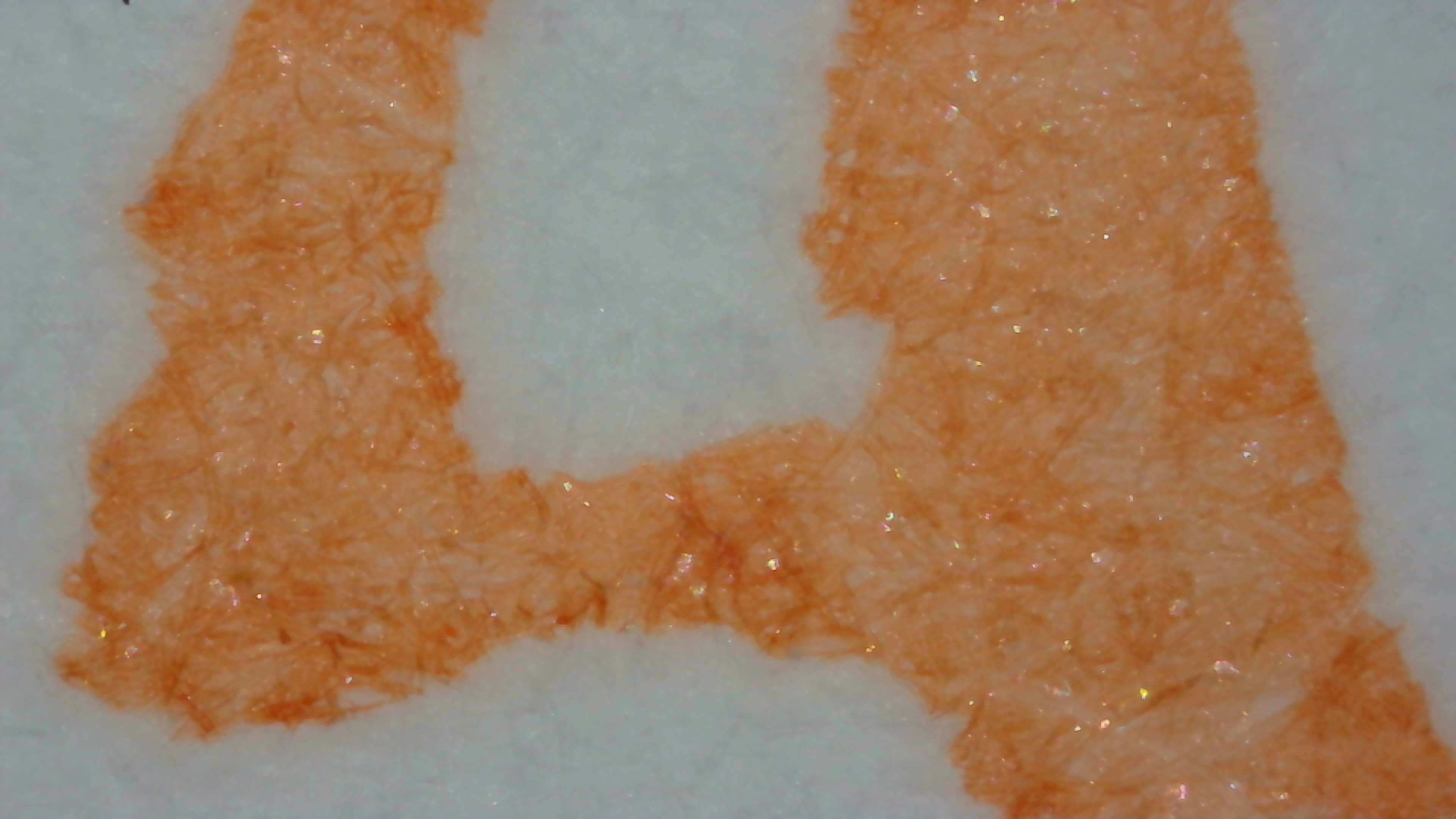 Photo of ink on paper under magnification on Col-o-ring paper. The ink is peach colored and very light. The edges of the lines are very rough, and a little darker in some places. There is a light amount of visible shimmer particles evenly distributed in the ink.