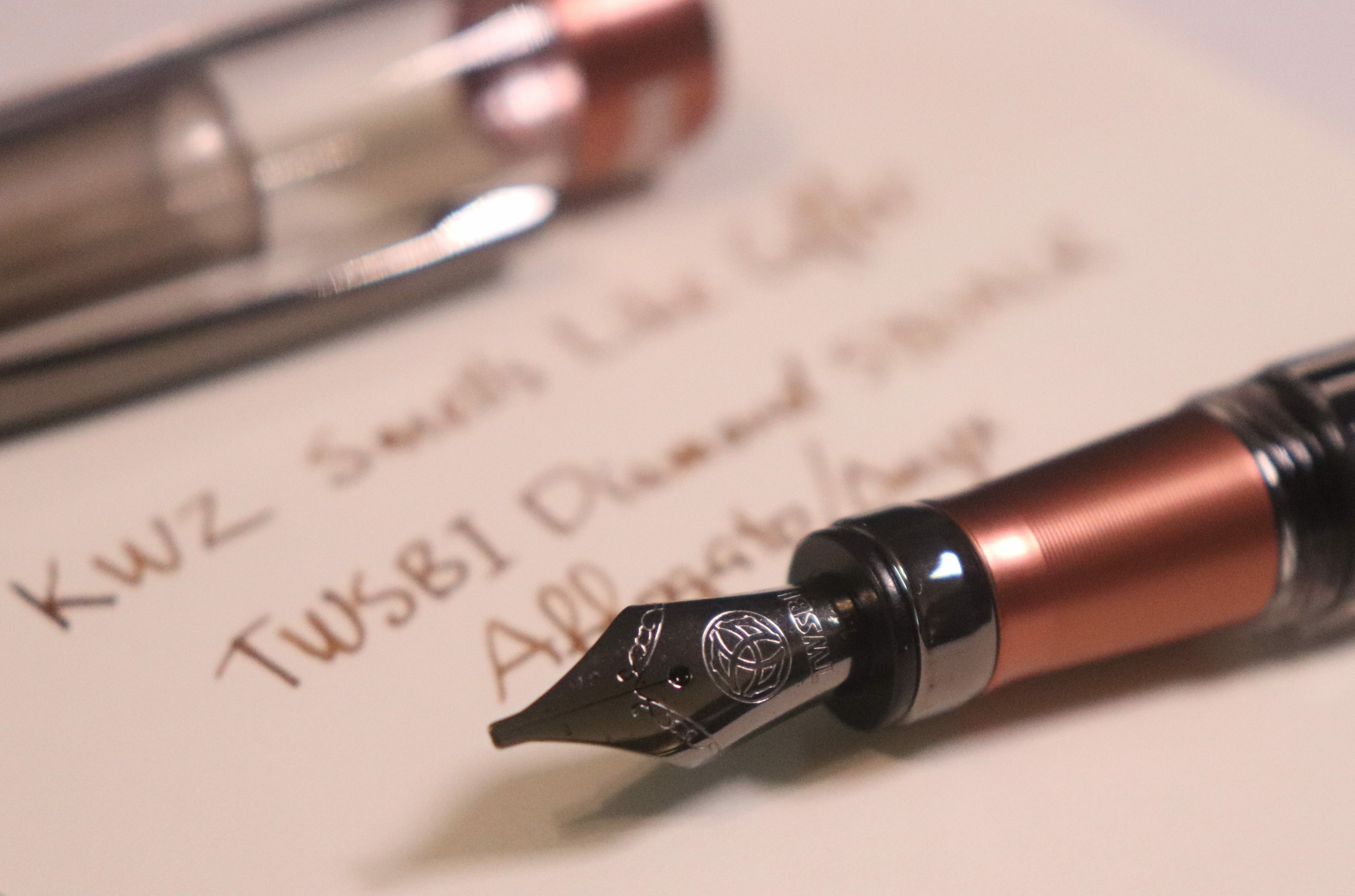 Photo of a fountain pen and writing sample on a piece of paper. The pen cap is removed and sitting across the top. The nib end of the pen is laying across the bottom. The pen is is clear plastic with metallic medium brown accents. The nib and clip are dark gray, almost black. Only the nib and front of the pen section are in focus, the writing sample is legible near the front but much blurrier in the toward the top of the photo. The writing sample reads "KWZ Smells Like Coffee / TWSBI Diamond 580ALR Affogato/Onyx" in medium brown ink.