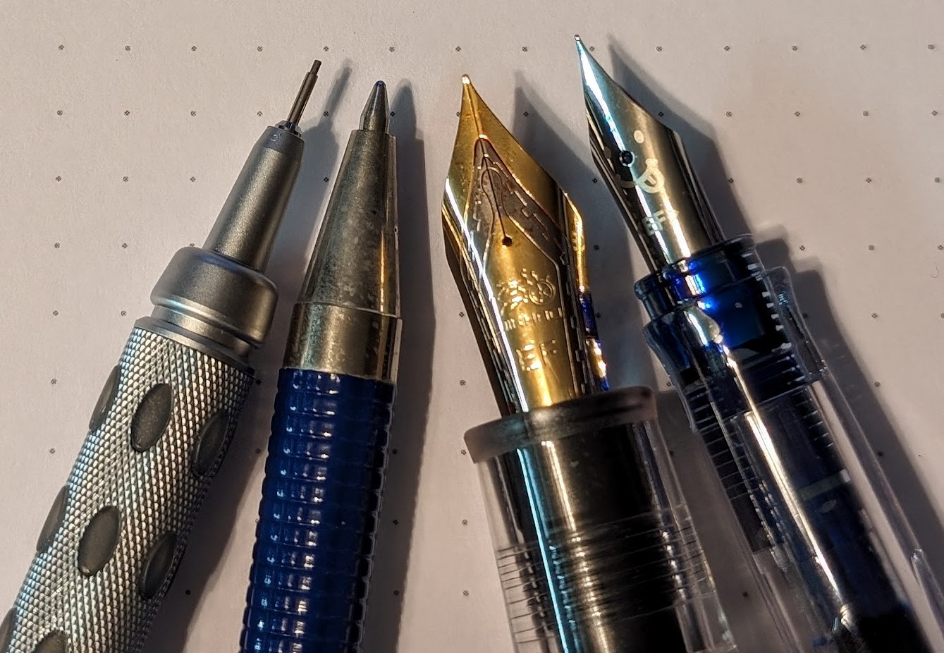 Photo of four writing implements arranged with their tips pointed toward the same point slightly out of frame. The focus is on the tip of each implement. They are, from Left to Right: * Pentel Graphgear 1000 PG1015 * Zebra F-301Rollerball * Jinhao 9019 Dadao #8 EF Nib * Pilot Kakuno EF Nib
