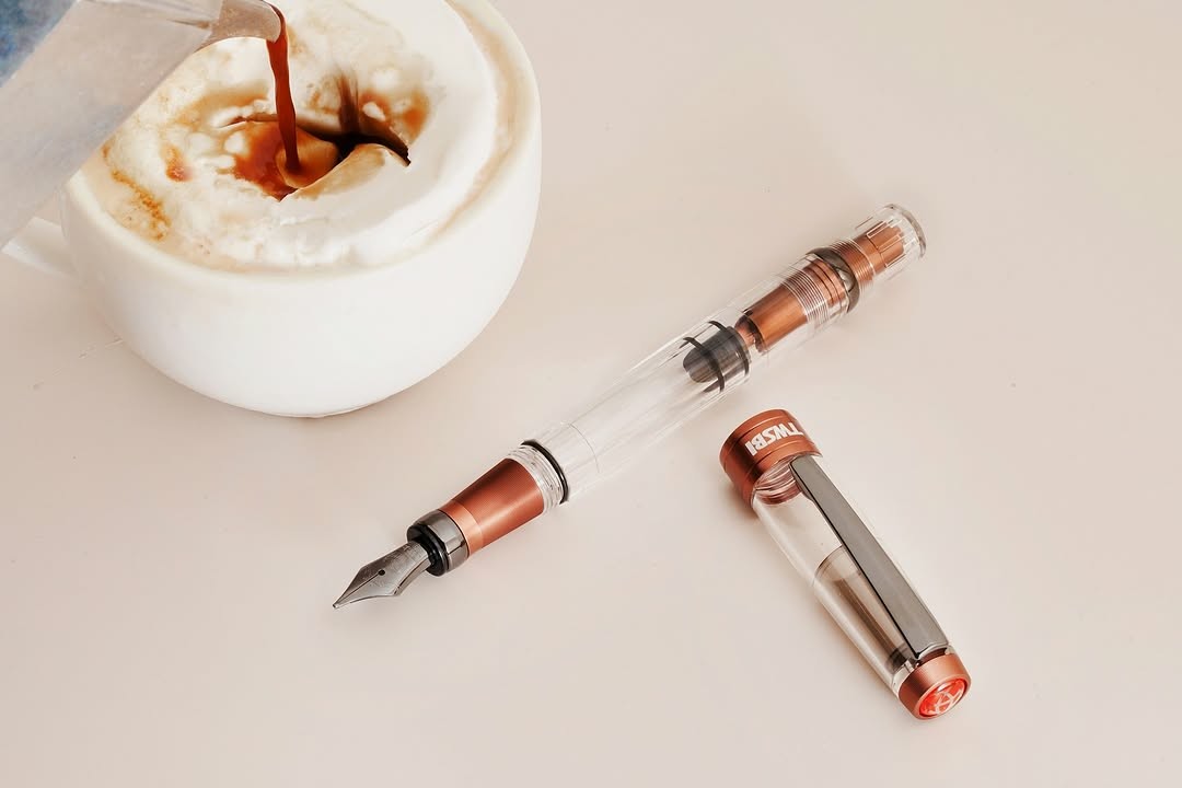 Photo of a fountain pen next to a cup with coffee and either gelato or ice cream (affogato) The pen has a clear body with brown metal about the same color as coffee around the grip section and inside the piston knob, with dark "onyx" trim and nib.