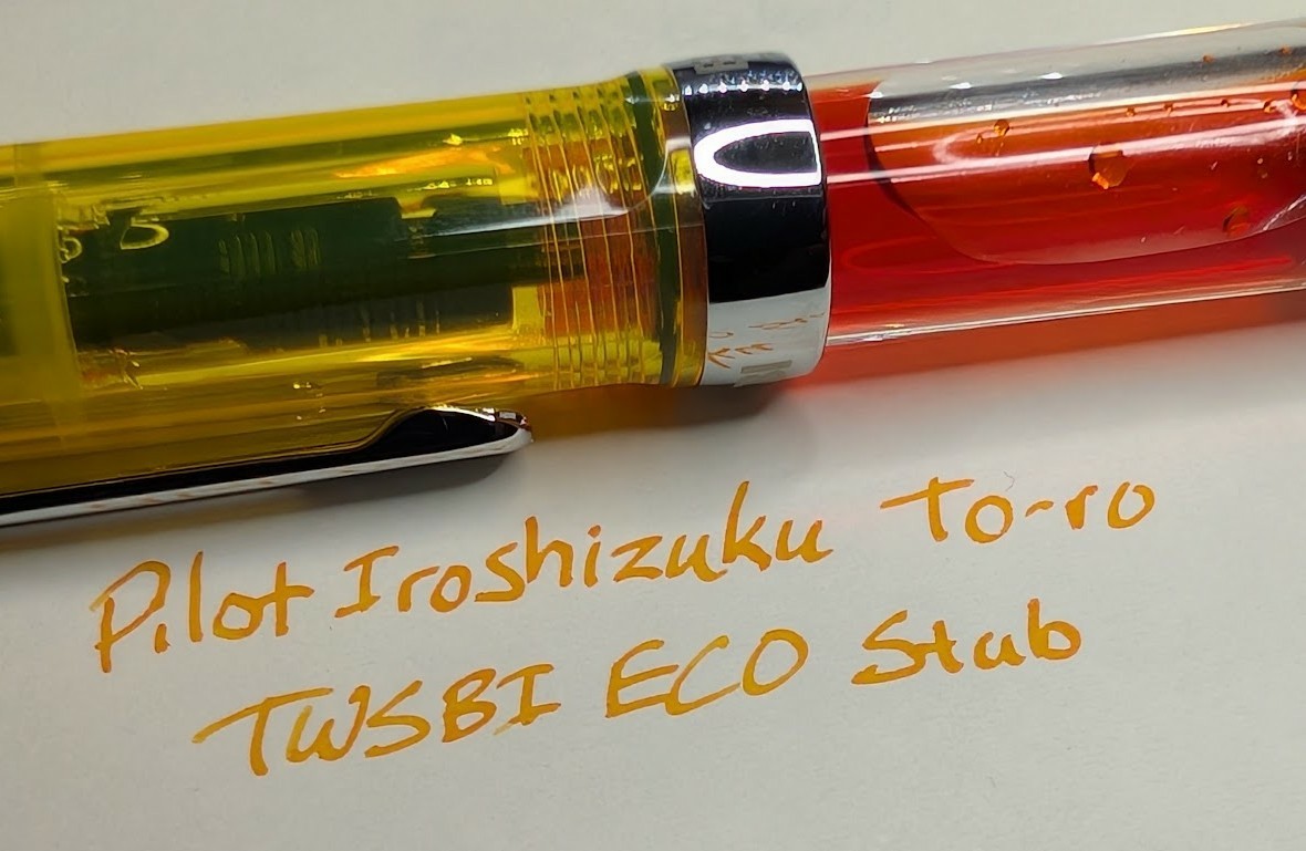 Photo of a fountain pen and writing sample on paper. The fountain pen is a TWSBI ECO with a transparent yellow cap. The accents and clip on the cap are silveer. The body of the pen is transparent and orange-yellow ink is visible inside. The writing sample reads "Pilot Iroshizuku To-ro / TWSBI ECO Stub" in yellow-orange ink.