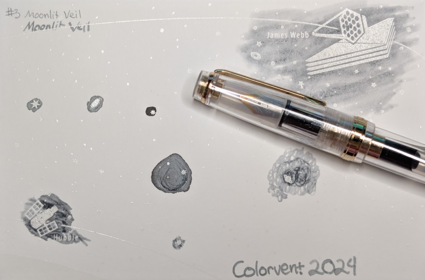 Photo of a "Colorverse Colorspace Size C" swatch card, type 1 which is based on the James Webb space telescope. The ink on the card is light gray. Upper left is text reading "#3 Moonlit Veil" written in an EF nib and then a stub nib. Below the text are two stars and a galaxy outlined in ink. Upper right is a large illustration of the James Webb telescope in negative space among swabbed ink. In the lower left is a small illustration of the Hubble space telescope in scribbled ink. In the middle lower left is a blob of ink with some visible lines where it dried darker. In the middle right is a blob of swirled ink drawn by a glass dip pen. Lower right is text "Colorvent 2024". Laying across the card is a Jinhao 82 fountain pen with a Fude nib. It is clear/transparent plastic with gold accents. Ink is visible in the section and converter inside the body.