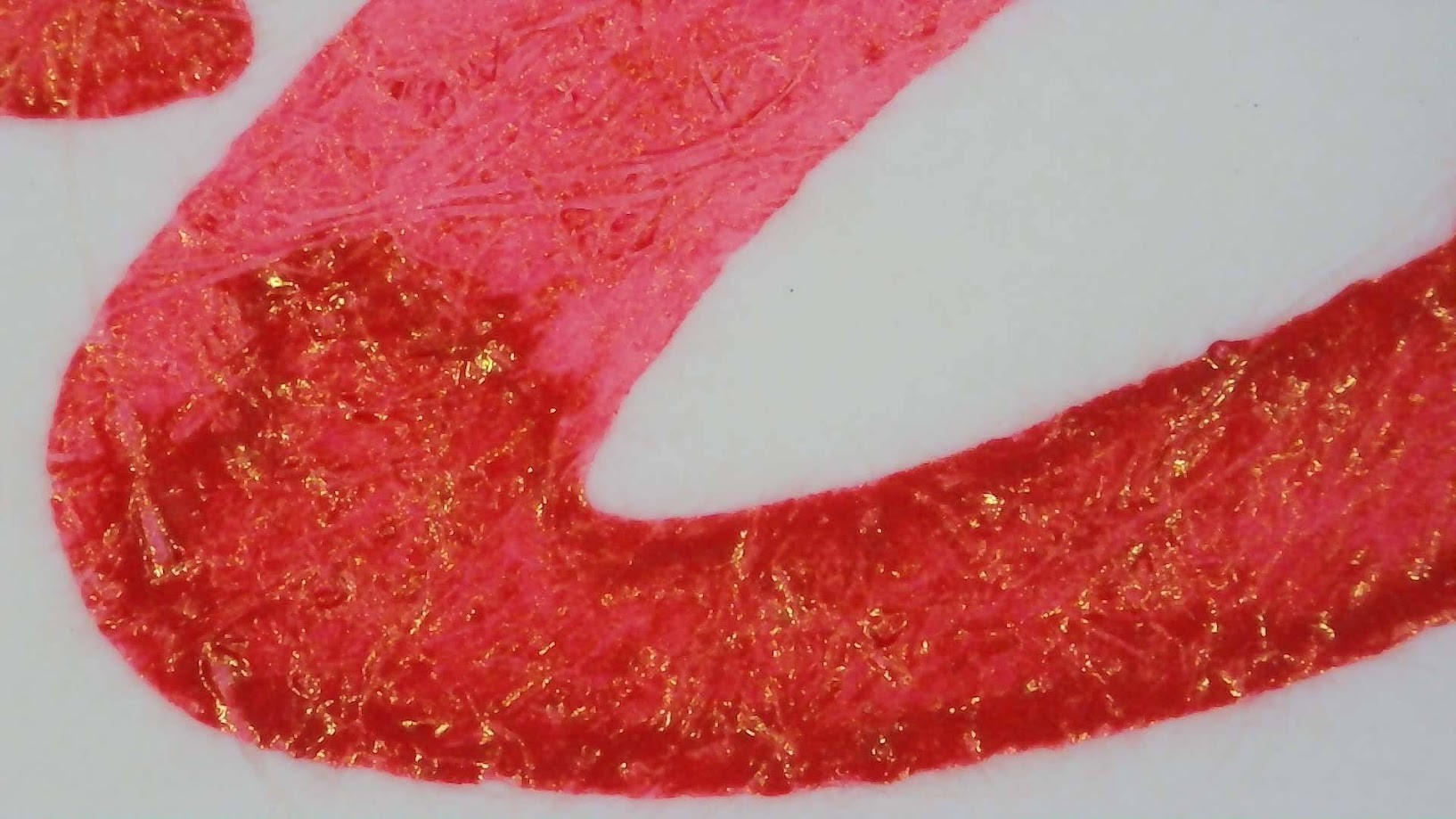 Microscope photo of ink on Iroful paper. The ink is bright red. In places with less ink it appears slightly pinkish. In places with more ink it's darker and the edge of those boundaries is darker still. Some flecks of gold are visible in the dark areas.
