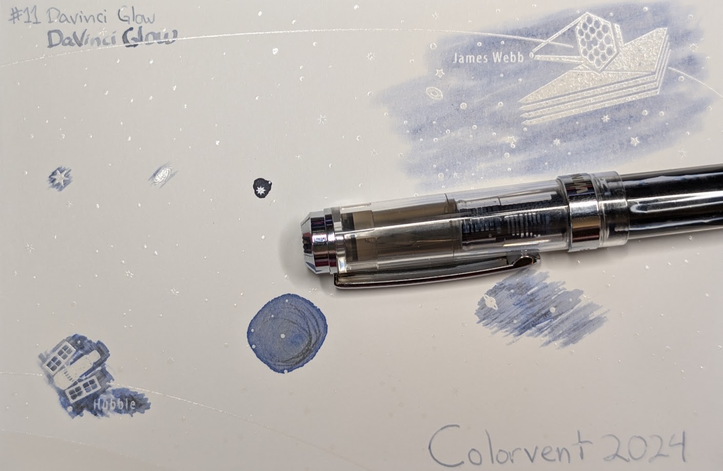 Photo of a "Colorverse Colorspace Size C" swatch card, type 1 which is based on the James Webb space telescope. The ink on the card is light blue-gray with some areas that are darker, and in the dark areas it is blue in places and dark gray in others. It is more blue near the edges and more gray in some areas with lots of ink. Upper left is text reading "#11 DaVinci Glow" written in an EF nib and then a stub nib. Below the text are multiple stars and a galaxy outlined in ink. Upper right is a large illustration of the James Webb telescope in negative space among swabbed ink. In the lower left is a small illustration of the Hubble space telescope in scribbled ink. In the middle lower left is a blob of ink with some visible areas where it dried darker. There are lines of gray in the center and a ring of blue around the outside. In the middle right is a blob of swirled ink drawn by a glass dip pen. Lower right is text "Colorvent 2024". Laying across the card is a fountain pen, a transparent WingSung 698 with silver accents.