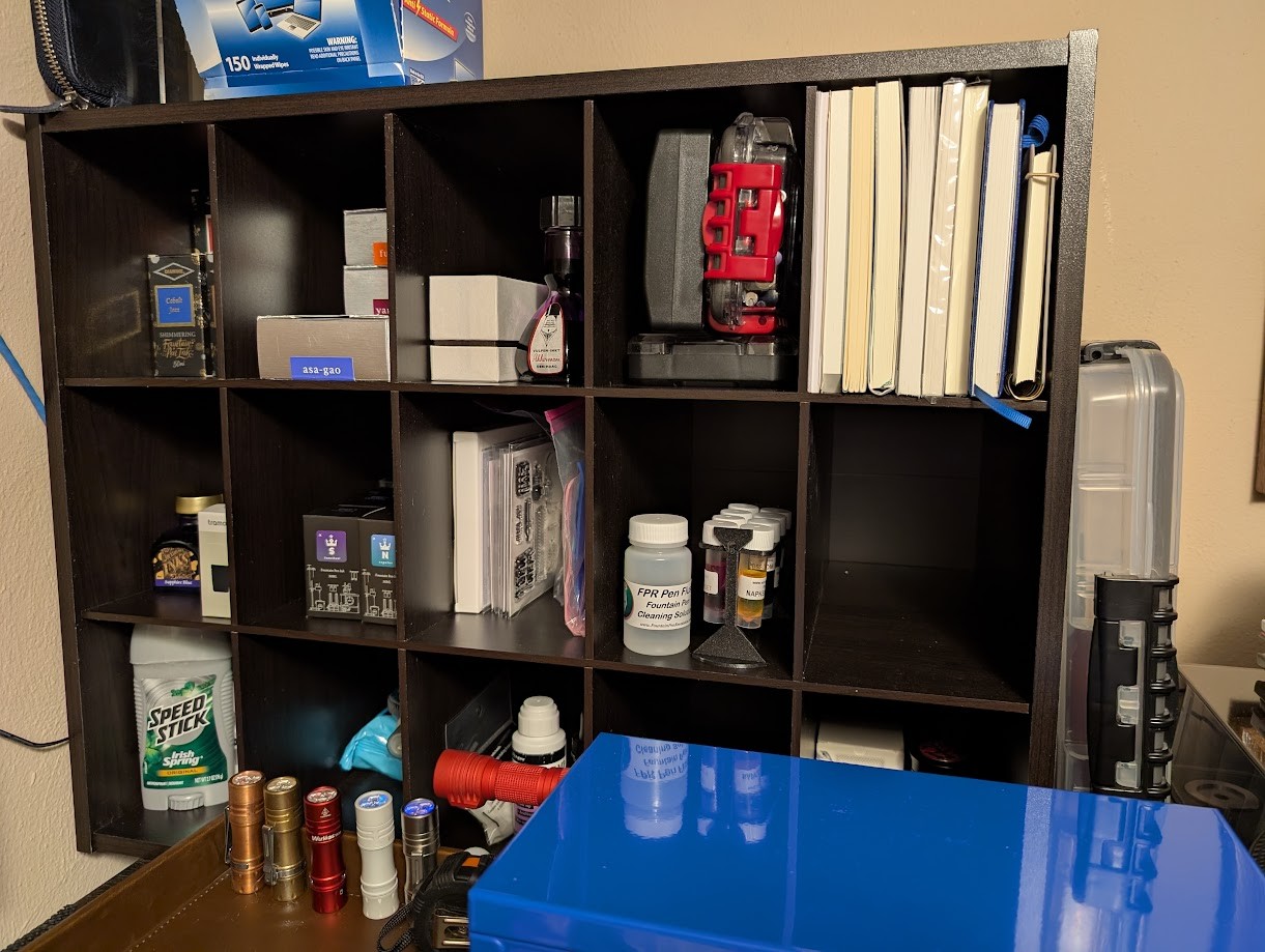 Photo of a "cubby" style rectangular shelf on top of a dresser. It is wider than it is tall. It is divided into 15 rectangular cubbies. In the cubbies are: Diamine ink, Pilot ink, more Pilot and Akkerman ink, tools and pens, several A5 notebooks that fit perfectly standing on their spine, and an A6 notebook, more Diamine and other misc ink, Tono & Lims ink, photopolymer stamps and ink, pen flush and samples, and some other personal care items (it is on top of a dresser, after all) On top is a container of electronics cleaning wipes and a leather folio. In front is a line of small flashlights and a metal "toolbox" style organizer.