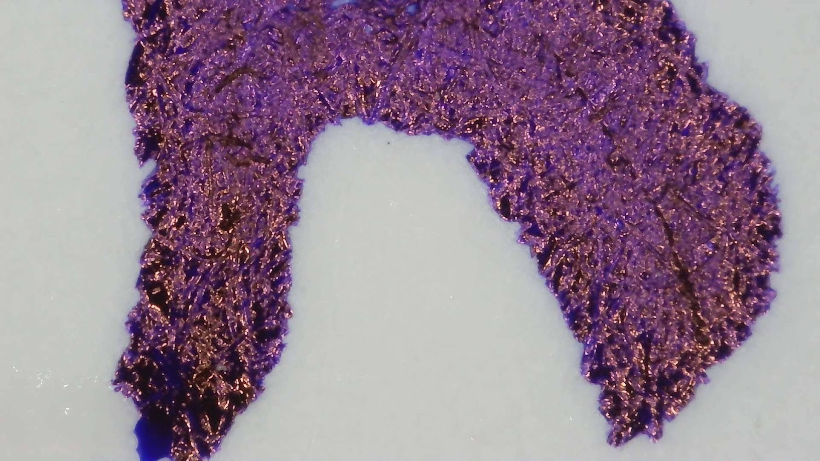 Photo of bluish purple ink writing under magnification. No letters can be discerned. Paper fibers are visible. Near the edge the ink is reflective and shiny red.