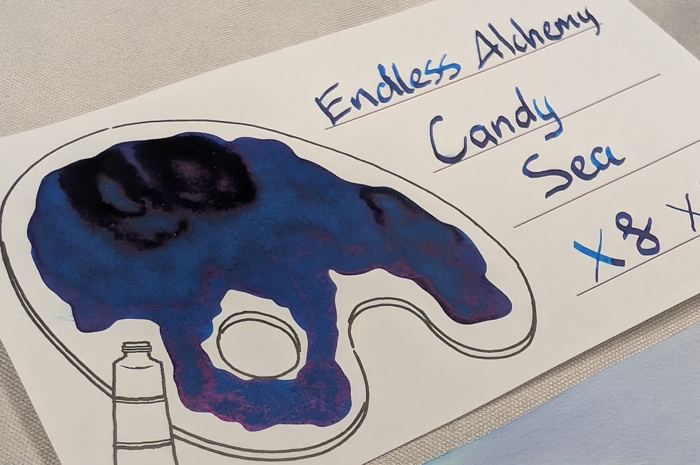 Even lower angle photo an ink swatch card. Overall the ink is a deep, dark blue with some areas of even darker blue, almost black. The card is an Iroful swatch card with an outline of a paint palette. Inside the palette is mostly filled in with swirls of midnight blue ink. Some areas with more ink are even darker blue, and areas with even more ink are almost black.