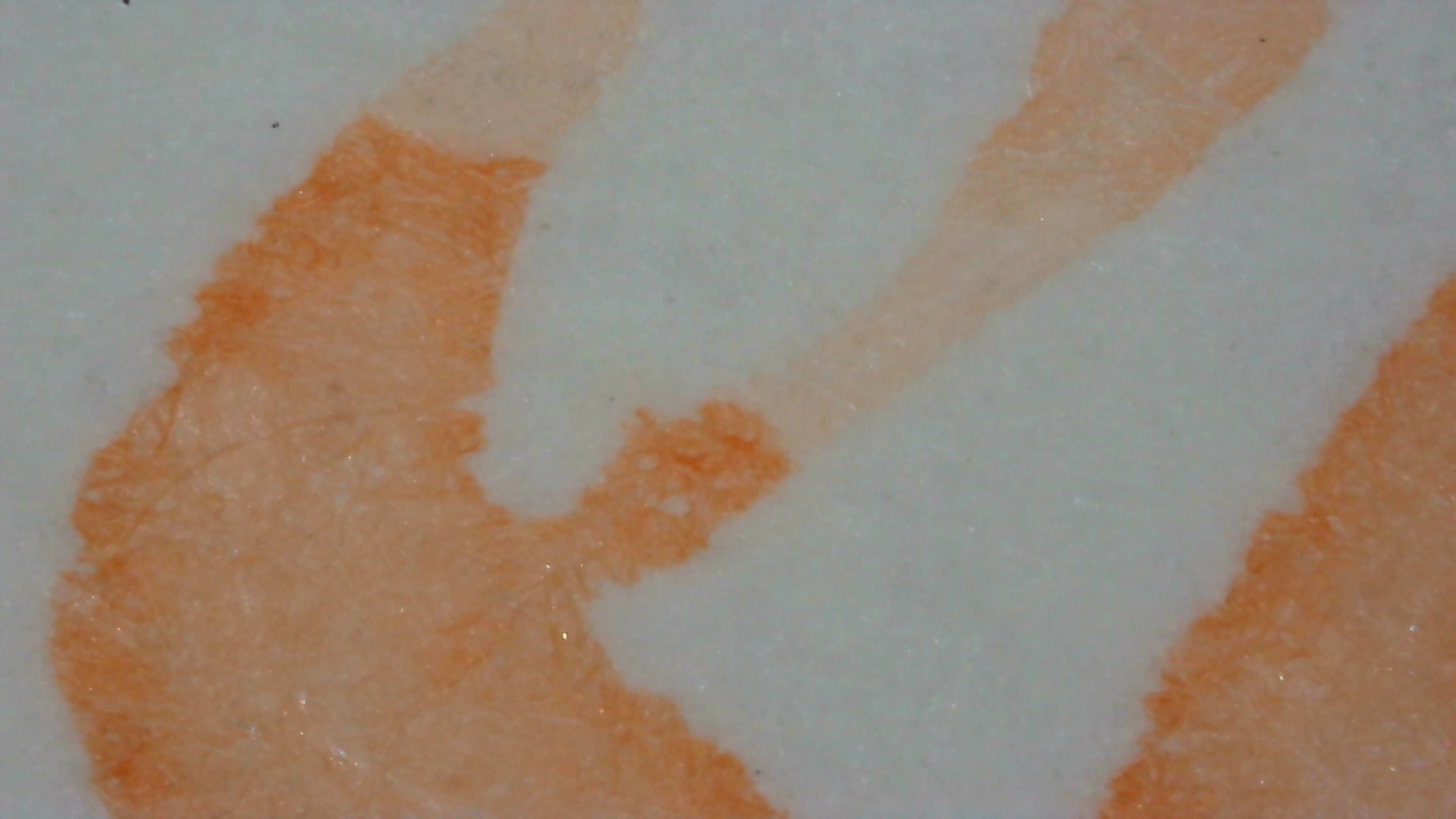 Photo of ink on paper under magnification on Midori paper. The ink is peach colored and very light. The edges of the lines are somewhat smooth, and on the left side they are a little darker. In the upper part of a loop in the line a section is even more pale. There is a light amount of visible shimmer particles evenly distributed in the ink.