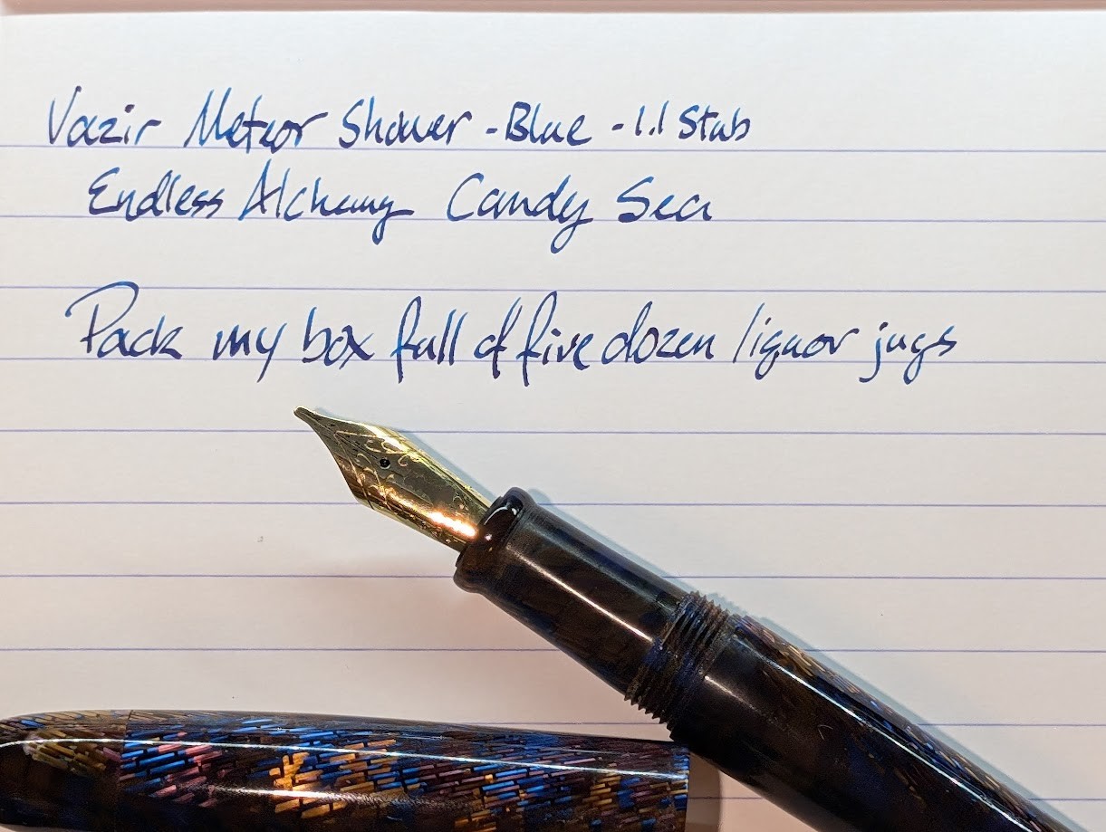 Photo of a writing sample which reads: "Vazir Meteor Shower - Blue - 1.1 stub Endless Alchemy Candy Sea Pack my box full of five dozen liquor jugs" Under the writing sample is the uncapped pen, sitting at an angle, resting against its cap.