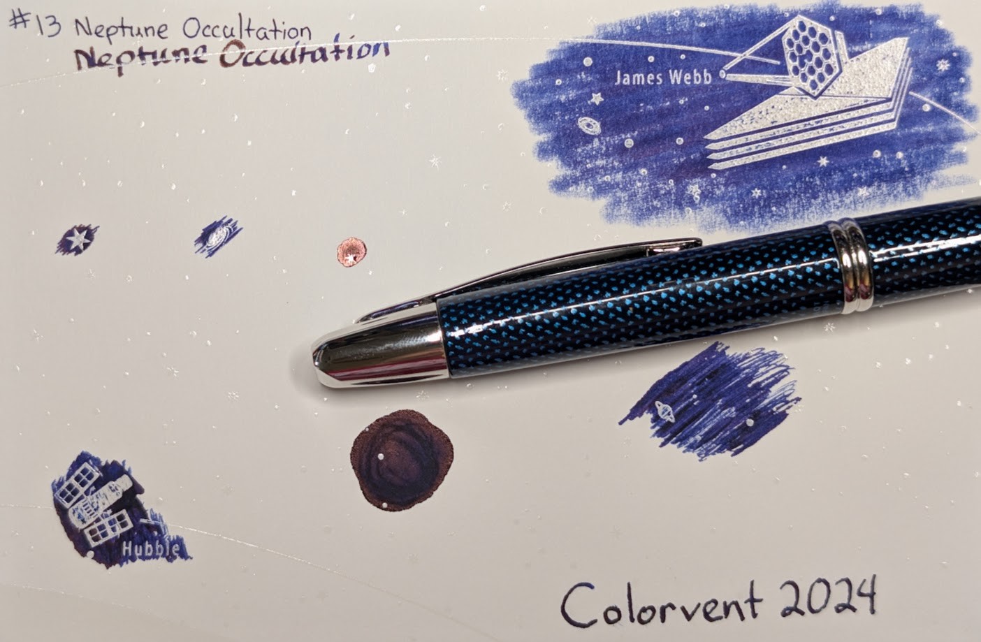 Photo of a "Colorverse Colorspace Size C" swatch card, type 1 which is based on the James Webb space telescope. The ink on the card is dark blue with some shading areas dark purple and some areas with coppery sheen. Upper left is text reading "#13 Neptune Occultation" written in an EF nib and then a stub nib. Below the text are multiple stars and a galaxy outlined in ink. Upper right is a large illustration of the James Webb telescope in negative space among swabbed ink. In the lower left is a small illustration of the Hubble space telescope in scribbled ink. In the middle lower left is a blob of ink with some visible areas where it dried darker and has coppery sheen. There are lines of dark purple in the center. In the middle right is a blob of scribbled ink drawn by a glass dip pen. Lower right is text "Colorvent 2024". Laying across the card is a fountain pen, a Pilot Vanishing Point (Blue Carbonesque) with a 1.1 stub nib.