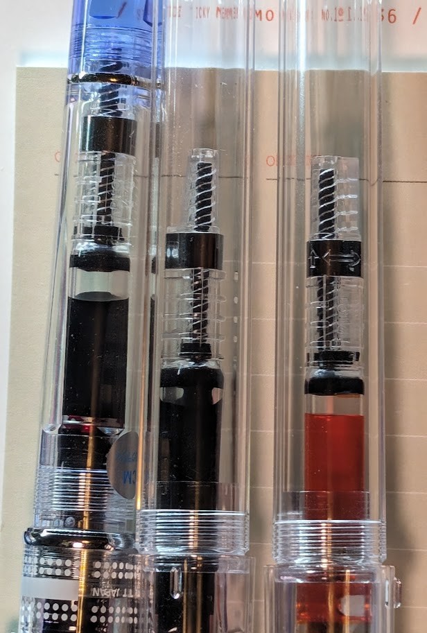 Three clear "demonstrator" fountain pens side-by-side: A Pilot Prera and two Pilot Kakunos. Only the main bodies of the pens are visible with a converter inside. Each of the three pens has a Pilot CON-40 converter inside which is almost completely full of ink. From left to right the ink colors are: - Pilot Iroshizuku Yama-Budo (Purple) - Diamine Writer's Blood (Dark Red) - Diamine Sunshine Yellow (Yellow with a slight orange tone)