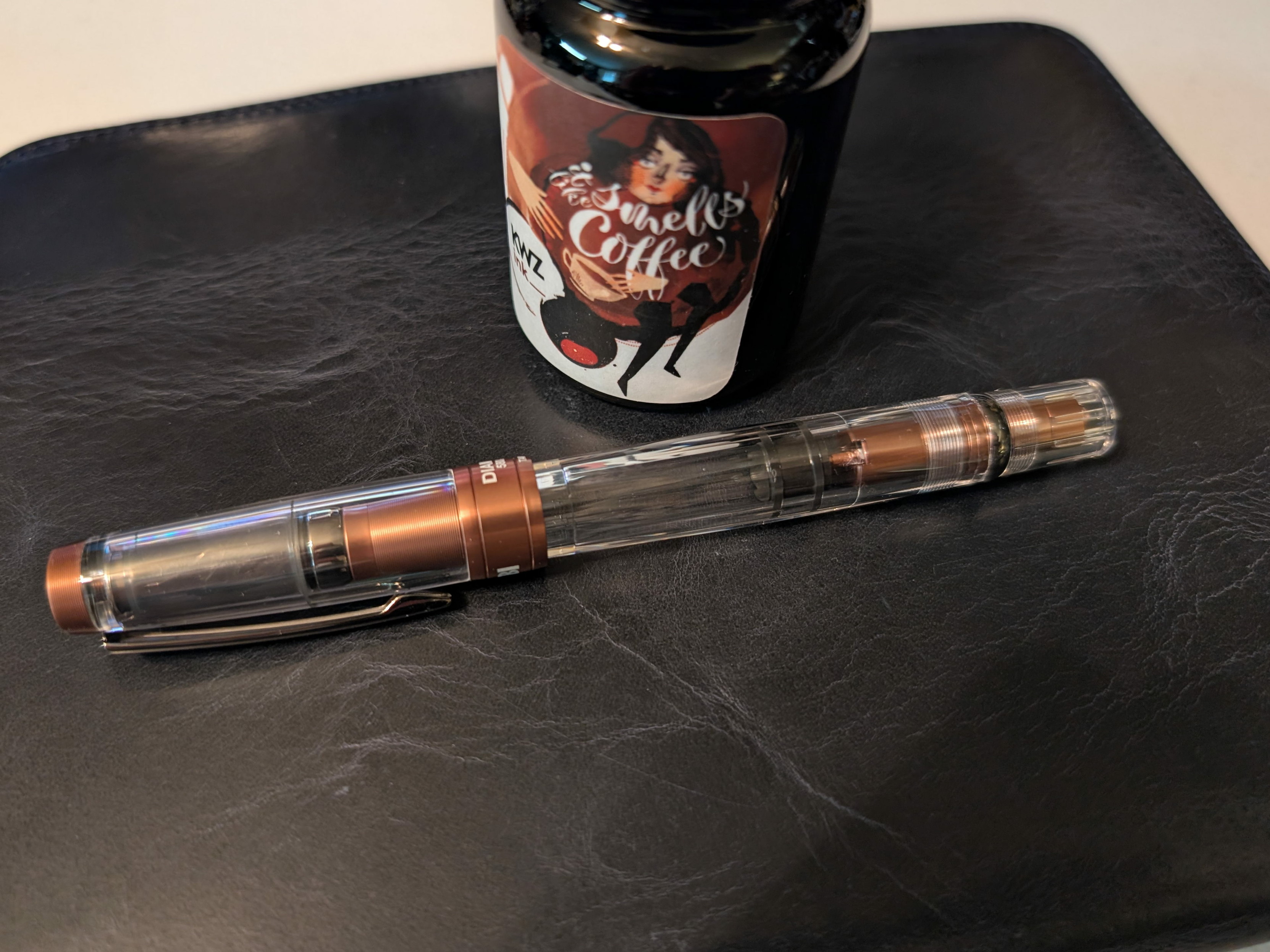 Photo of a fountain pen and bottle of ink on a dark navy blue folio. The pen is laying in front of the ink bottle. The pen is is clear plastic with metallic medium brown accents. The clip is dark gray, almost black. The bottle of ink is labeled "KWZ It Smells Like Coffee" and has a sketch of a woman holding a coffee cup, the name of the ink is in white between the woman's face and the coffee cup.