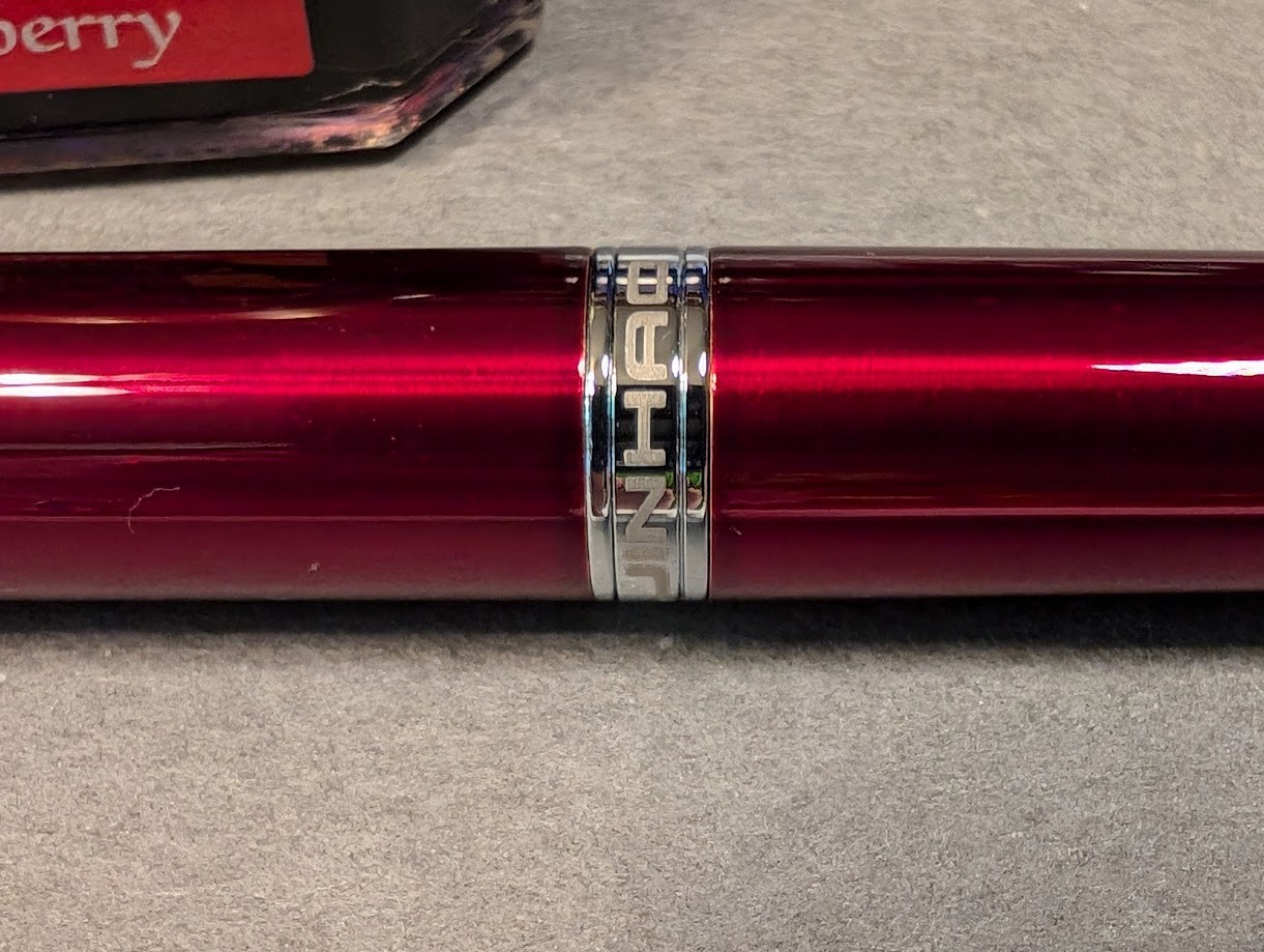 Photo of a fountain pen next to a bottle of ink, close-up. The bottle of ink is square-ish and the only visible text reads "berry". The fountain pen is dark red and shiny with a silver ring approximately in its center. In the ring is some writing: "JINHAO" in all capital letters.