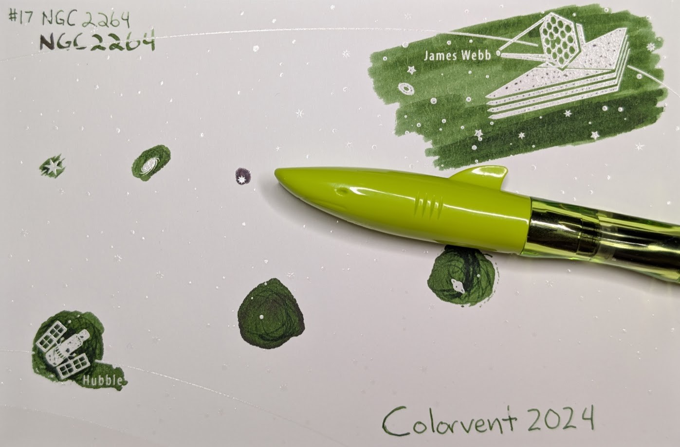 Photo of a "Colorverse Colorspace Size C" swatch card, type 1 which is based on the James Webb space telescope. The ink on the card is medium forest green with some shading. Upper left is text reading "#17 NGC 2264" written in an EF nib and then a stub nib. Below the text are multiple stars and a galaxy outlined in ink. Upper right is a large illustration of the James Webb telescope in negative space among swabbed ink. In the lower left is a small illustration of the Hubble space telescope in scribbled ink. In the middle lower left is a blob of ink with some visible areas where it dried darker and has reddish sheen. There are lines of darker ink in the center. In the middle right is a blob of scribbled ink drawn by a glass dip pen. Lower right is text "Colorvent 2024". Laying across the card is a green Jinhao Shark fountain pen with a transparent green section. The cap of the pen is shaped like the front of a shark, with a mouth, eyes, and a fin.