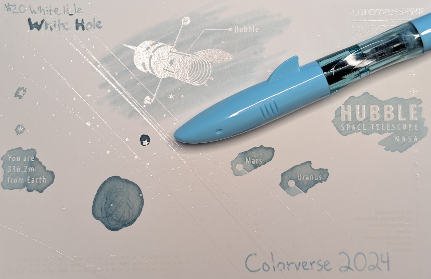 Photo of a "Colorverse Colorspace Size C" swatch card, type 2 which is based on the Hubble space telescope. The ink on the card is a very pale light blue with some darker areas were more ink settled on the paper. Upper left is text reading "#20 White Hole" written in an EF nib and then a stub nib. Some letters are extremely light, others have areas with much darker ink. Just to the left side of the middle at the top is a large illustration of the Hubble space telescope in negative space among swabbed ink. In the middle right side is text "Hubble Space Telescope NASA" in negative space among scribbled ink. In the middle left is text reading "You are 336.2mi from Earth" in negative space among ink scribbles. In the middle lower left is a blob of swirled ink drawn by a glass dip pen. Near the center are "Mars" and "Uranus" with circles representing those planets, in more swabbed ink. Lower right is text "Colorverse 2024" and the ink is a much more even shade that is brighter blue, not as pale. Laying across the card is a light blue Jinhao Shark fountain pen with a transparent light blue section. The cap of the pen is shaped like the front of a shark, with a mouth, eyes, and a fin.