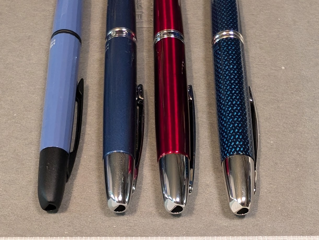 Photo of four fountain pens, placed vertically and partially visible. Photo is focused on the tips of the pens. From left to right they are: * Majohn A2 which is a flat blue-gray color with black trim and a faceted surface. * Majohn A1 in a dark shiny metallic blue color with silver trim and smooth surface. * Jinhao 10 Press in dark shiny red color with silver trim. * Pilot Vanishing Point in Blue Carbonesque pattern which is dark blue with a repeating pattern of lighter blue hashed-like splotches. The surface is slightly textured. It has silver trim. The tip of all four pens looks eerily similar, but there are slight variations in the rings and clips.