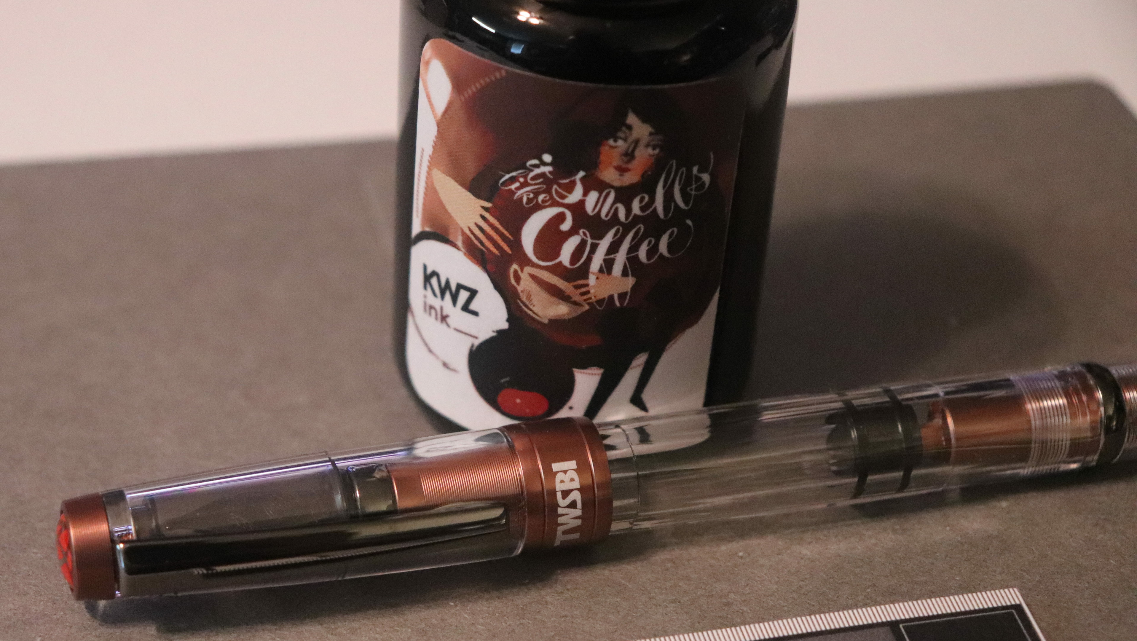 Photo of a fountain pen and bottle of ink on a dark gray notebook. The pen is laying in front of the ink bottle. The pen is is clear plastic with metallic medium brown accents. The clip is dark gray, almost black. The bottle of ink is labeled "KWZ It Smells Like Coffee" and has a sketch of a woman holding a coffee cup, the name of the ink is in white between the woman's face and the coffee cup.