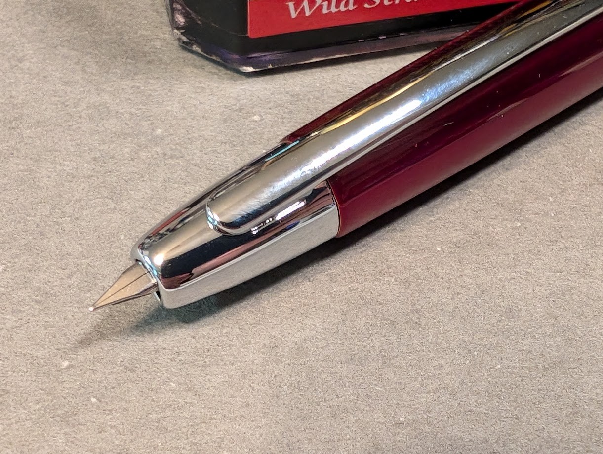 Photo of a fountain pen next to a bottle of ink. The bottle of ink is square-ish and partially visible, the visible text reads "Wild S". The fountain pen is dark red and shiny with a silver tip and silver clip. The pen is capless and resembles a Pilot Vanishing Point. The nib is extended and visible, coming to a sharp point at the tip.