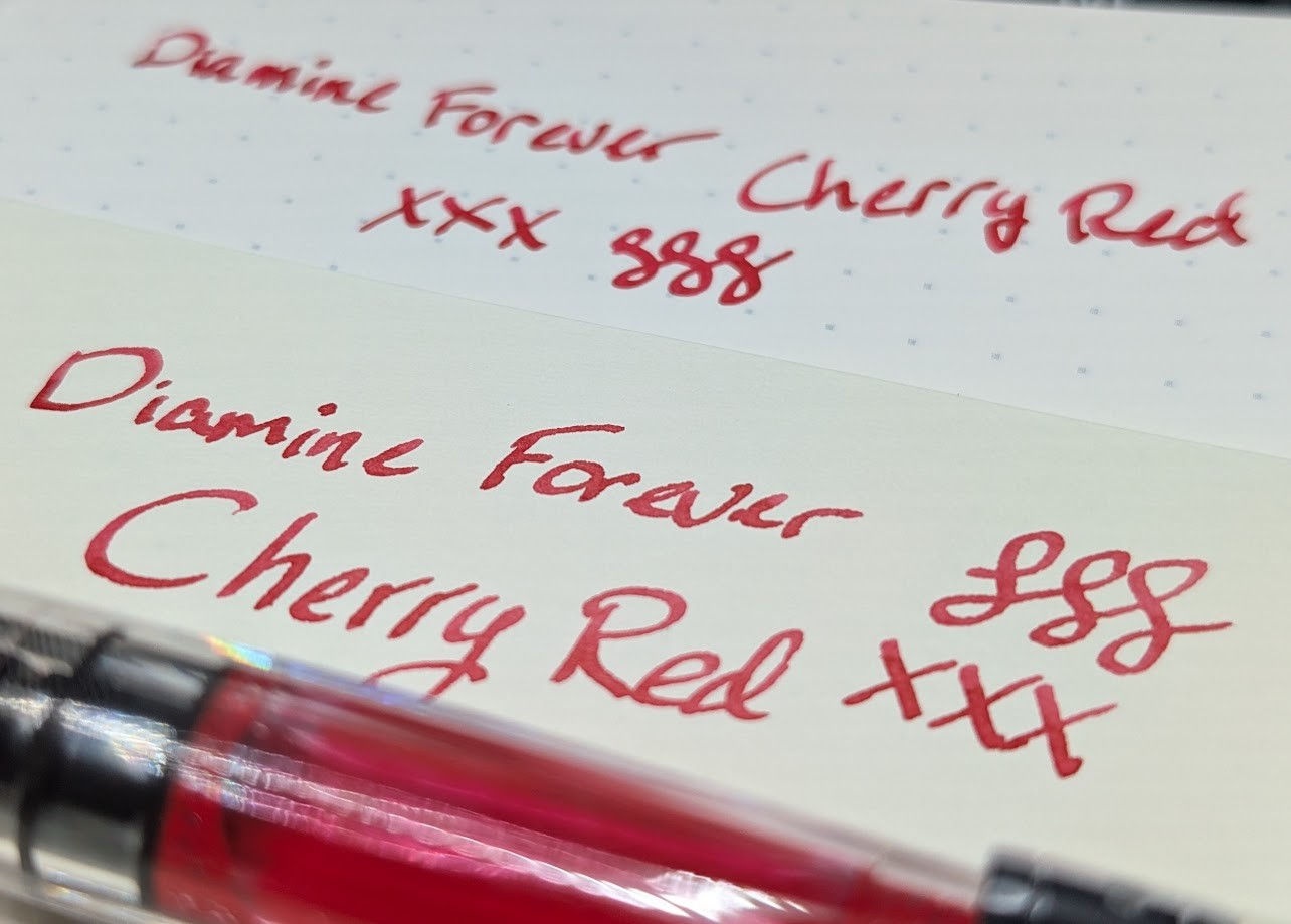 Photo of two writing samples and a fountain pen containing red ink. Taken from a low corner angle. Both samples say "Diamine Forever Cherry Red" followed by three "X"s and three curlicues. The ink is a bold red.
