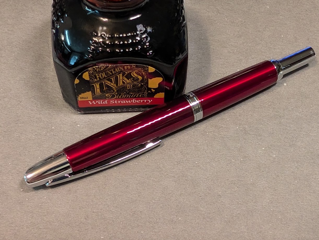 Photo of a fountain pen next to a bottle of ink. The bottle of ink is square-ish and reads "Fountain Pen Inks by Diamine" and "Wild Strawberry". The fountain pen is dark red and shiny with a silver tip, silver clip, silver ring approximately halfway down the body, and a silver tail end. The pen is capless and resembles a Pilot Vanishing Point. The nib is retracted and not visible.