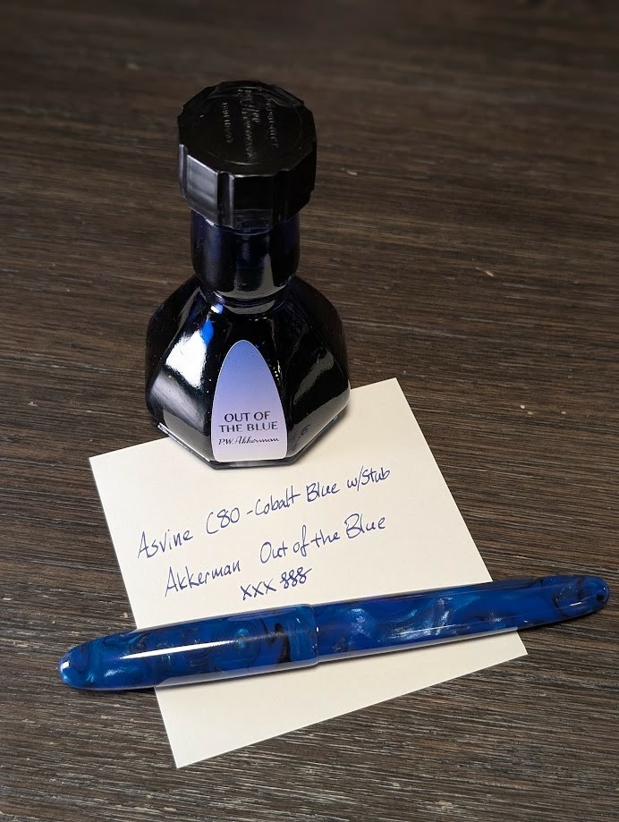 Photo of a fountain pen, ink bottle, and note paper with a writing sample all on a wooden table. The fountain pen is an Asvine C80 in Cobalt Blue, which is generally blue but has swirls of black, light blue, and shimmer all through the body and cap. The ink bottle is octagonal at the base, then rises up to a narrow "waist" then changes to a cylinder at the top which extends up several cm to the cap. The writing sample reads "Asvine C80 - Cobalt Blue w/Stub / Akkerman Out of the Blue" followed by several "X" characters and curlicues -- The ink is a shade of blue which is close to the pen.