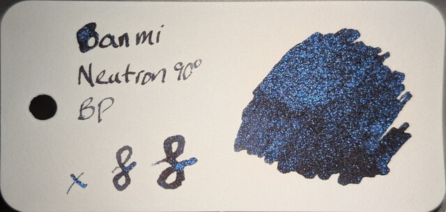 Photo of an ink swatch on a col-o-ring swatch card.  Text on the left reads "Ban Mi Neutron 90° BP" with an "x" and two loops next to it. On the right is a large ink blob made with a glass dip pen.  The base ink is dark, black or extremely dark gray. The ink contains many tiny reflective particles of blue glitter which catch the light.