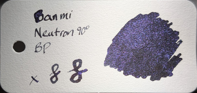 Photo of an ink swatch on a col-o-ring swatch card.  Text on the left reads "Ban Mi Neutron 90° BP" with an "x" and two loops next to it. On the right is a large ink blob made with a glass dip pen.  The base ink is dark, black or extremely dark gray. The ink contains many tiny reflective particles of purple glitter which catch the light.